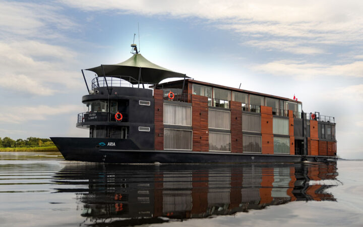 Aria Amazon ship with 2 decks, tall cabin windows, black metal & warm wood, cruises a calm river under a partly cloudy sky.