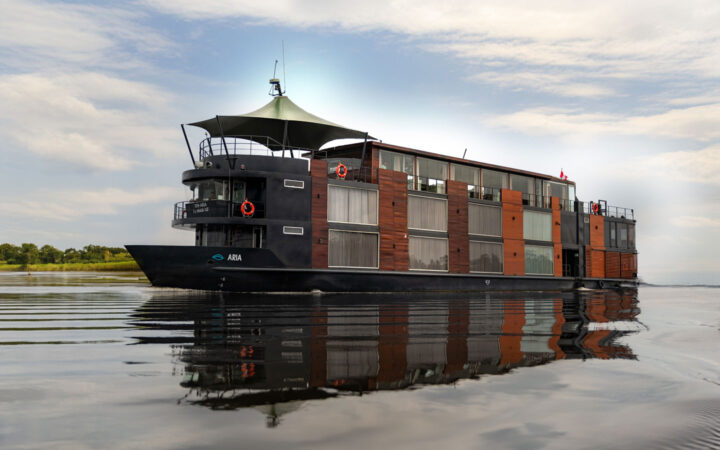 Aria Amazon ship with 2 decks, tall cabin windows, black metal & warm wood, cruises a calm river under a partly cloudy sky.