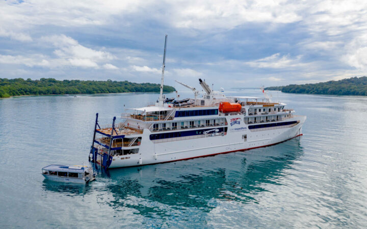Aerial view of Coral Adventurer ship with multiple guest decks, white exterior, hydraulic aft deck & Xplorer tender vessel.