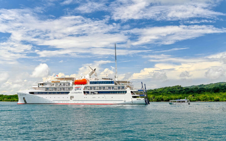 Exterior port side of Coral Geographer Australian expedition ship with gray metal tender vessel behind under blue sky.