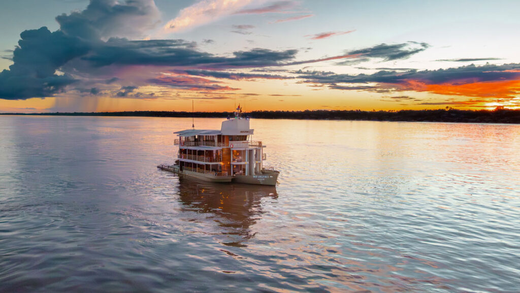 Aerial view of Delfin I Peruvian Amazon riverboat cruising in calm water at dusk under orange, pink, purple & blue sky.