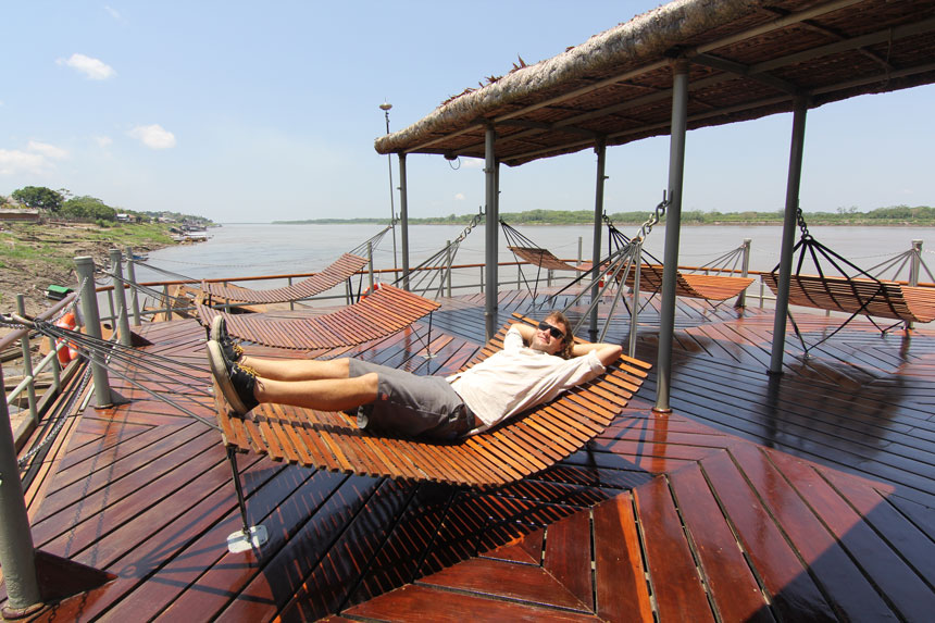 Man in light shirt & dark shorts lays on a wooden slatted hammock on top deck of Delfin I Amazon river ship on a sunny day.