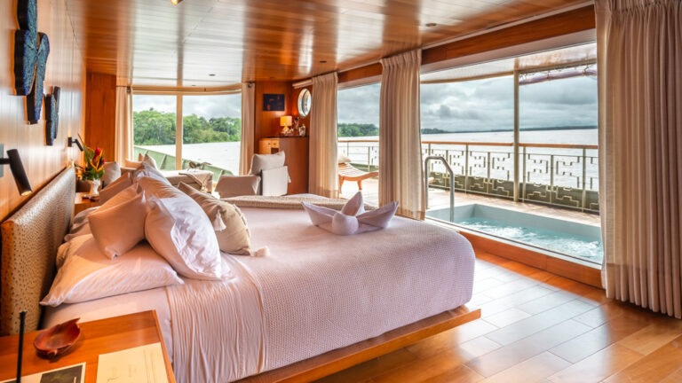 Deluxe bedroom aboard Delfin I with a double bed with white sheets, large floor-to-ceiling windows, and a small hot tub on the deck