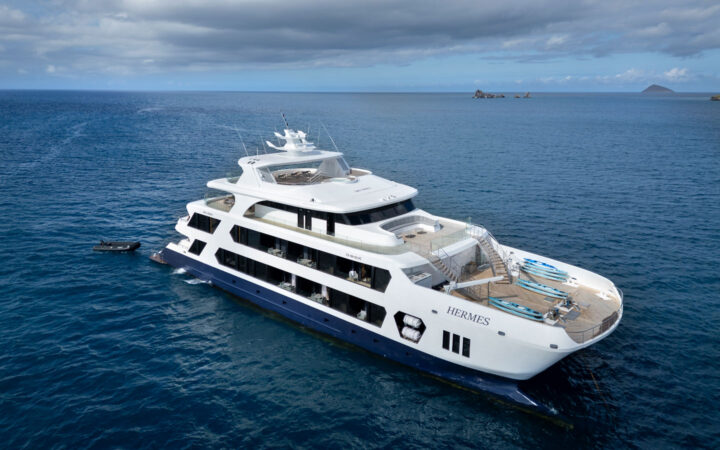 Aerial view of Hermes Galapagos luxury ship with 4 guest decks in white & blue with teak decking, sitting in calm sea.