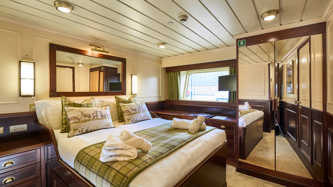A double bed with white sheets and 2 folded towels on a small cruise ship with brown interior.
