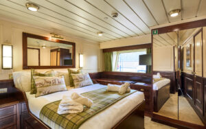 A double bed with white sheets and 2 folded towels on a small cruise ship with brown interior.