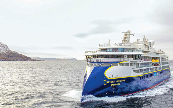 Sleek blue & white National Geographic Endurance expedition ship with multiple decks cruises through pale water on a gray day.