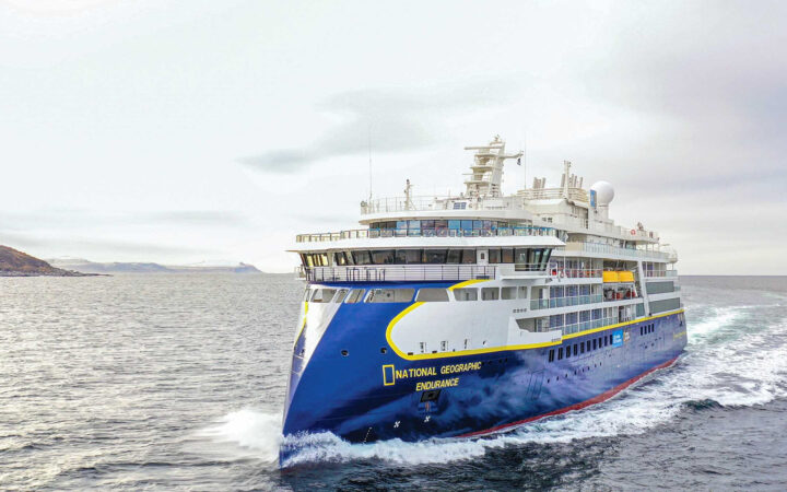Sleek blue & white National Geographic Endurance expedition ship with multiple decks cruises through pale water on a gray day.