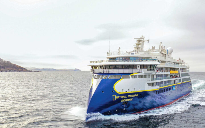 Sleek blue & white National Geographic Endurance expedition ship with multiple decks cruises through pale water on a gray day.