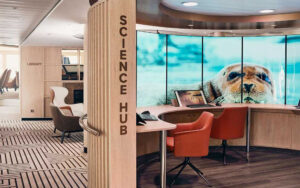 Science Hub of Nat Geo Endurance & Resolution with large wood desk, baby seal on TV screens & lab equipment.