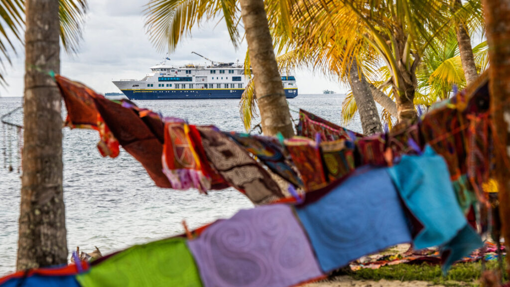 Colorful, handwoven squares hung on lines between palm trees in foreground & small blue, gold & white Nat Geo ship in background.