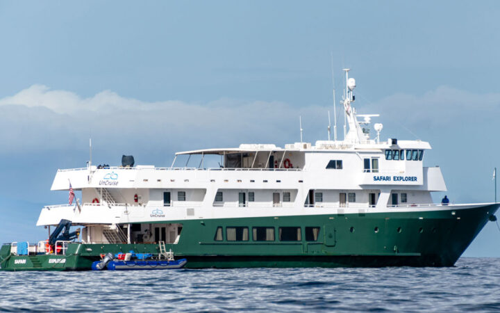 Small ship with green hull, UnCruise logo & Safari Explorer written on the side sits in calm water on a partly cloudy day.