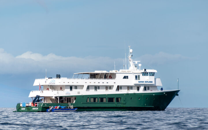 Small ship with green hull, UnCruise logo & Safari Explorer written on the side sits in calm water on a partly cloudy day.