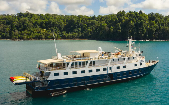 Small ship Safari Voyager with dark bue hull & white upper decks on anchor in tropical warters offshore from lush rainforest.