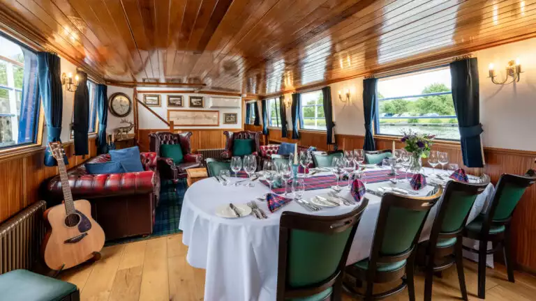Dining room table set for a meal with white-&-blue linens & wooden bar behind aboard Scottish Highlander small riverboat.