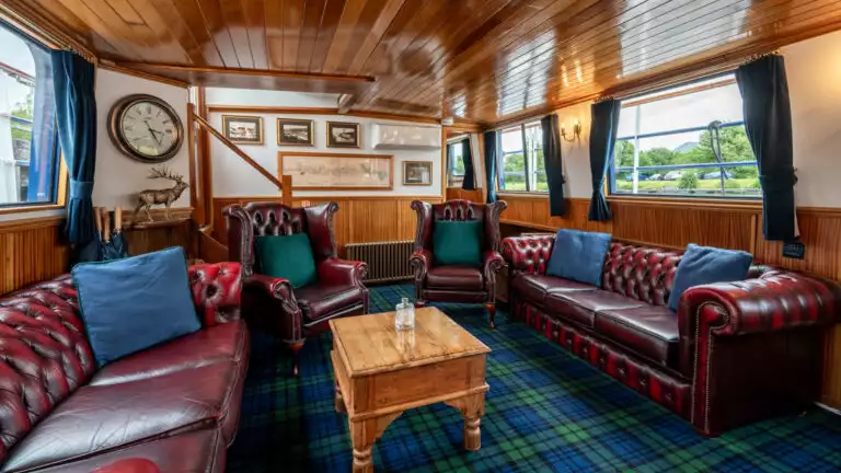 Saloon indoor common area with dining table set for service & living room behind with leather armchairs & sofas aboard Scottish Highlander Caledonia Canal small ship.