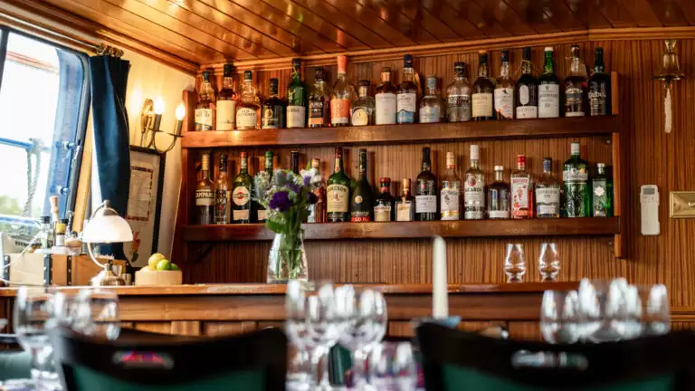 Warm wooden bar with 2 shelves of whisky behind & view window with vase of flowers beside it, aboard Scottish Highlander hotel barge.