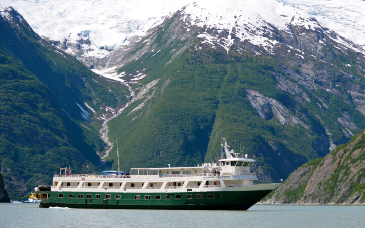 Wilderness Discoverer boat with forest green hull & 2 white upper decks cruises past green mountains with snow-covered peaks.