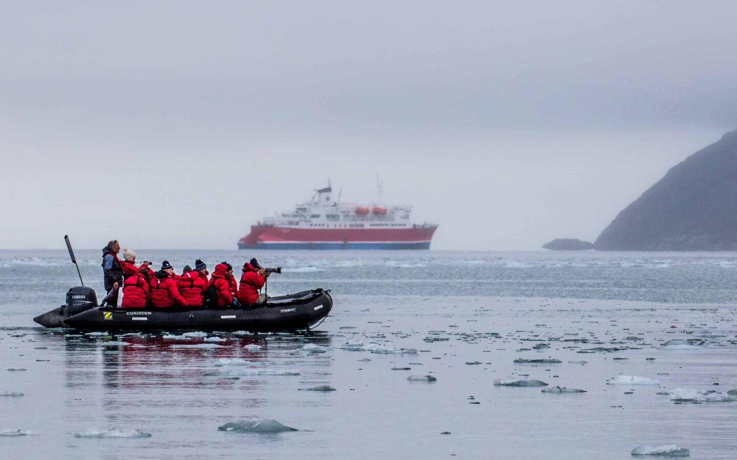 arctic cruise travelers in a zodiac skiff wearing red jackets look at the landscape with an expedition ship behind them