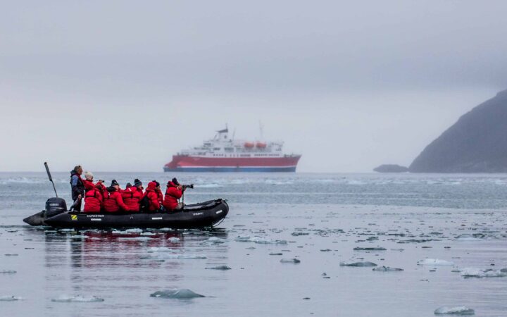 arctic cruise travelers in a zodiac skiff wearing red jackets look at the landscape with an expedition ship behind them