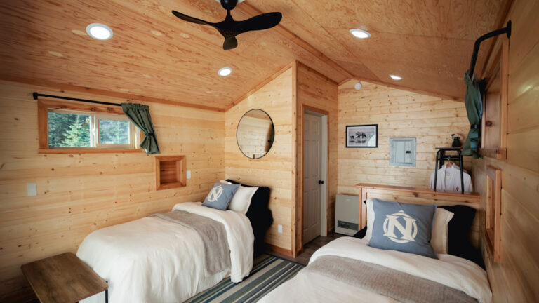 Wood cabin with two twin beds, tan bedding and grey pillows, with a fan and small windows