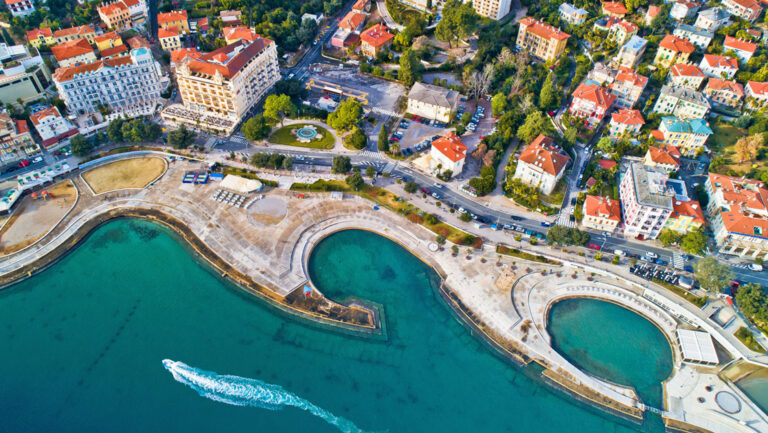 Aerial view of yacht cruising turquoise sea along riverfront of Opatija, Croatia with terracotta roofs & beige buildings.