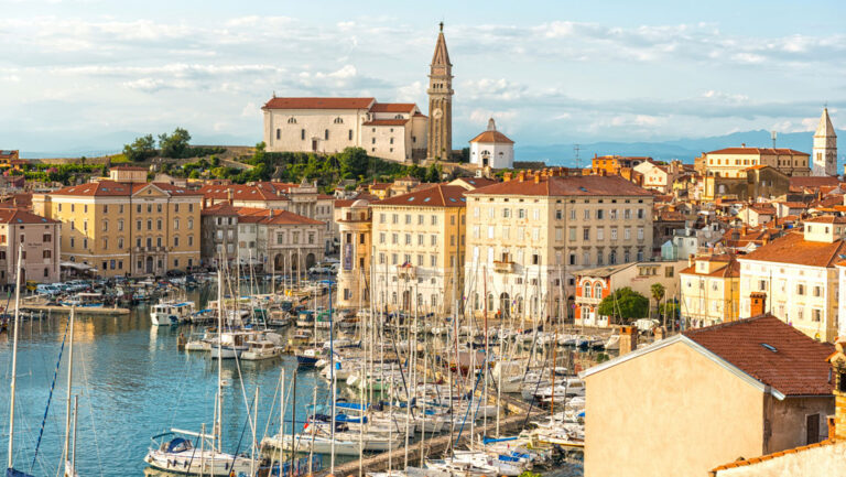 Clocktower of Piran, Slovenia rises above red-roofed beige buildings beside small harbor with sailboats & yachts docked.