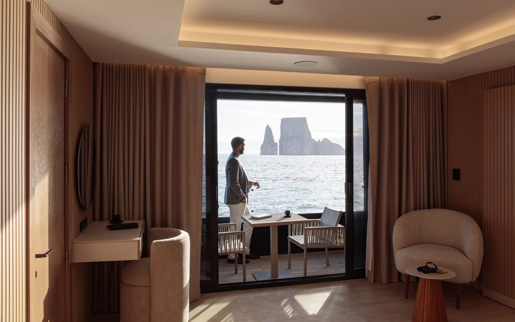 A man in a sports coat stands on the balcony of a luxury Galapagos cruise ship in front of Kicker Rock