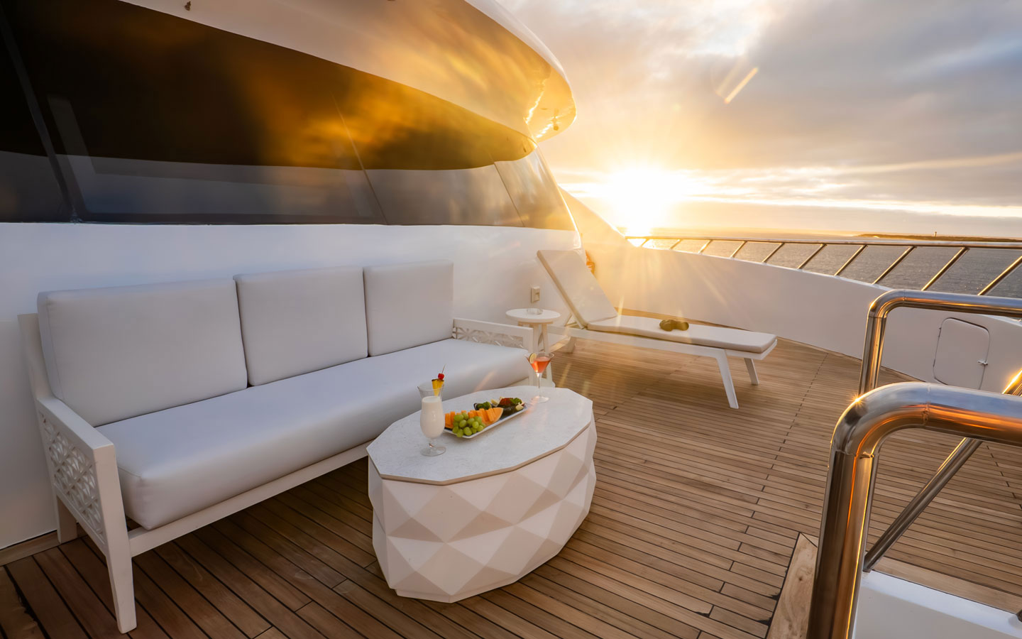 The wood deck bow of a Galapagos yacht charter seen with white couch, sun lounge chairs & coffee table set with cocktails & fruit at sunset.