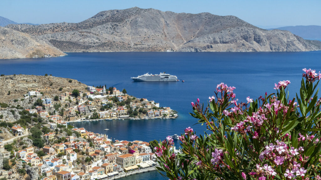 Aerial view of pink flowers overlooking seaside Greek island town with small gray expedition ship sitting offshore in the sun.