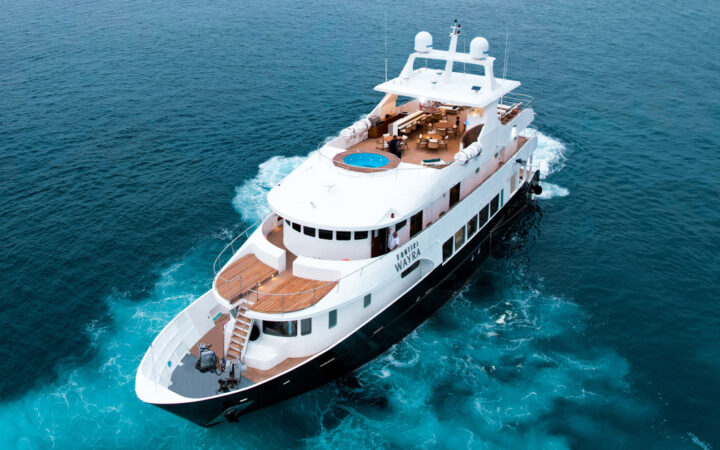 Aerial view of Kontiki Wayra yacht with dark blue hull, 3 white upper decks, teak decking & large Sundeck Jacuzzi.