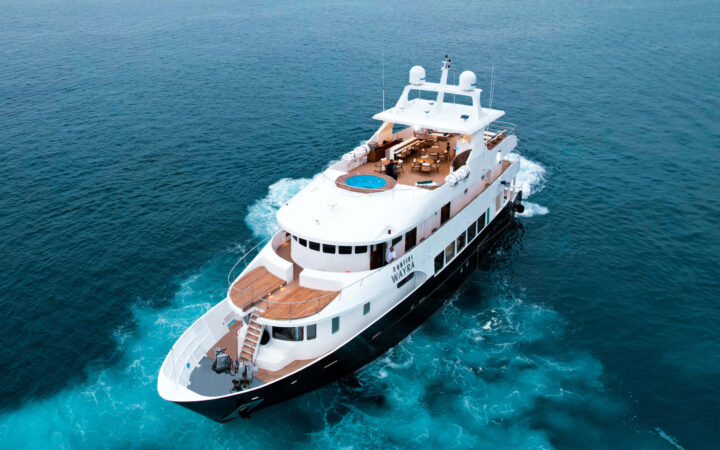 Aerial view of Kontiki Wayra yacht with dark blue hull, 3 white upper decks, teak decking & large Sundeck Jacuzzi.