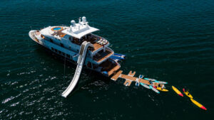 Aerial view of Kontiki yacht with blue hull, white & teak upper decks, waterslide, floating aft dock, paddleboards & kayaks.