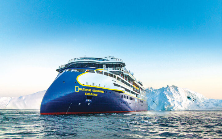 National Geographic Endurance ship with blue hull & sharp bow, operated by Lindblad Expeditions, sits in calm water by iceberg.