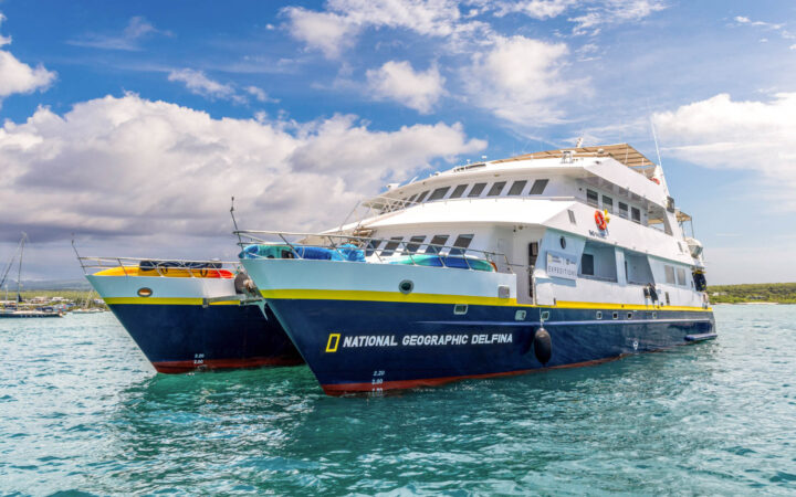 Luxury catamaran National Geographic Delfina painted white with a royal blue hull floats in the Galapagos on a partly sunny day