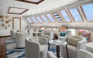National Geographic Delfina ship's lounge with windows, sunlight, white padded sofa bench & chairs & ethnic art.