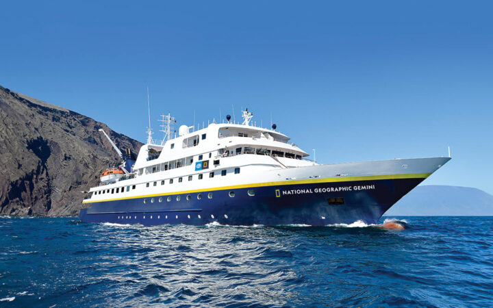 Exterior of Nat Geo Gemini, painted white with navy blue hull and floating in the ocean in blue sky day in Galapagos.