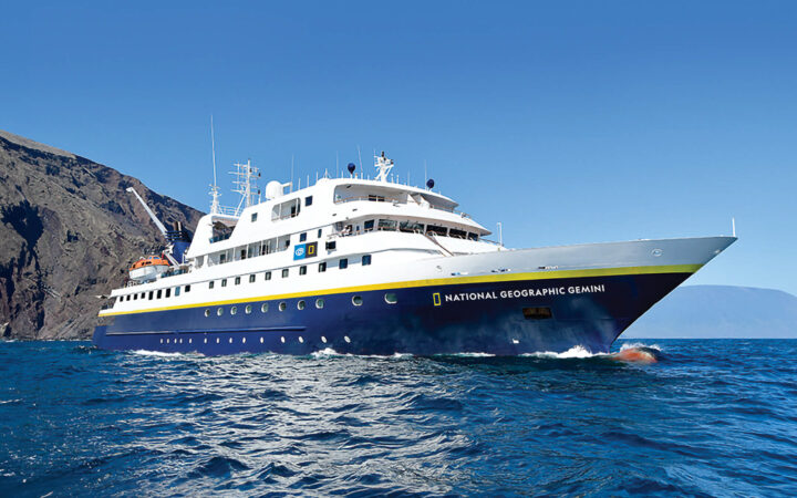 Exterior of Nat Geo Gemini, painted white with navy blue hull and floating in the ocean in blue sky day in Galapagos.