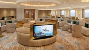Beige interior longue aboard Nat Geo Gemini. Upholstered accent chairs with picture windows and flat screen TV in the center.