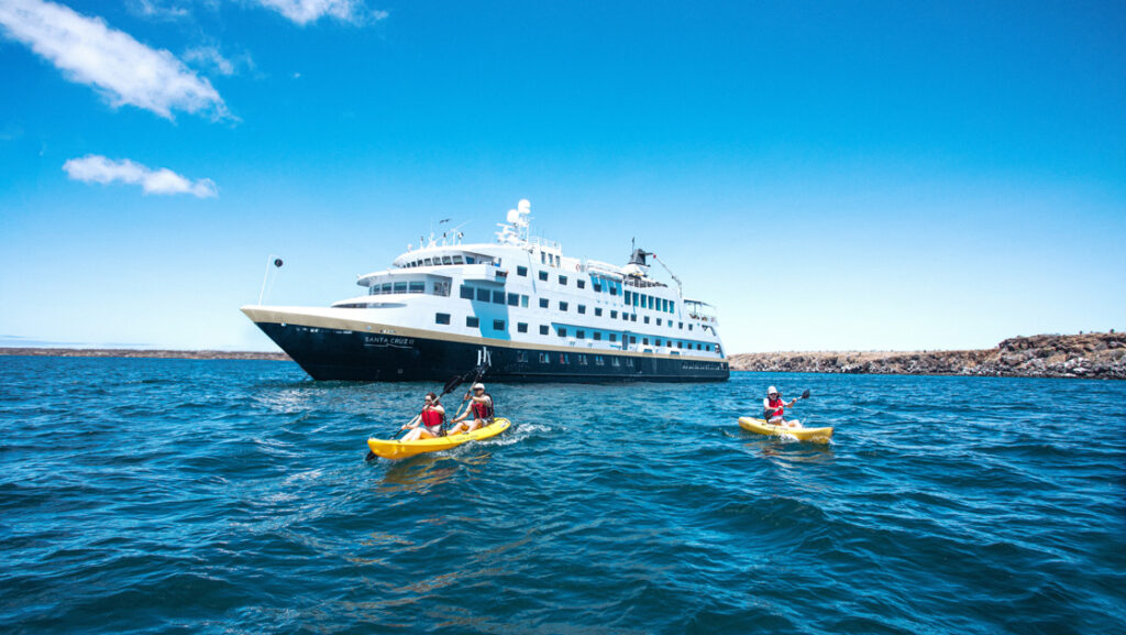 Santa Cruz II Galapagos ship with dark blue hull & gold & white upper decks sits in calm sea as guests paddle kayaks nearby.