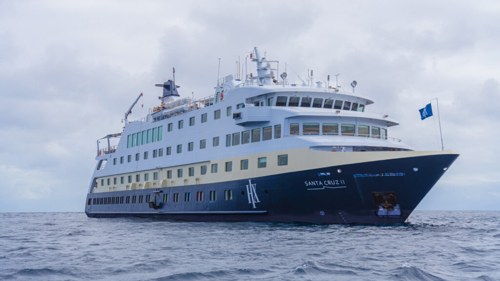 Santa Cruz II Galapagos small ship with dark blue hull & gold & white upper decks cruising on calm waters on a cloudy day.