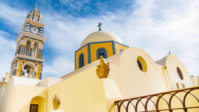 Yellow Byzantine architecture with domes & clocktower under blue sky on a sunny day in Santorini, Greece.