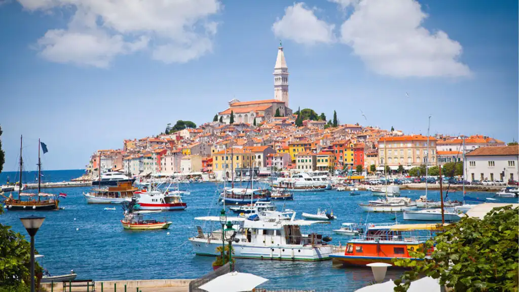 Busy Mediterranean harbor with yachts & fishing boats in blue sea by terracotta-roofed beige stone buildings in the Adriatic.