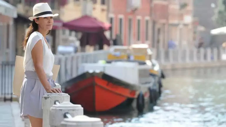 Woman in white hat & blouse & gray shorts stands along Venice's Grand Canal with a small riverboat nearby & colorful buildings.