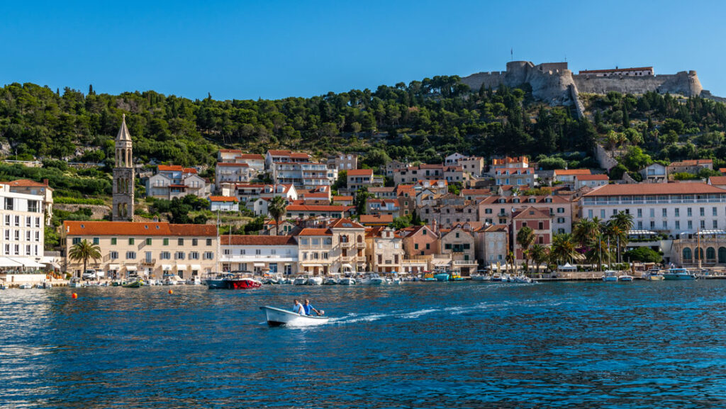 Seaside town in Croatia with terra cotta roofs on beige brick buildings on a green hillside topped by castle walls in the sun.