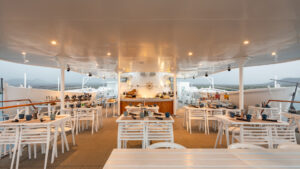 Dining area aboard small cruise ship with white tables and chairs and a bar in the back for guests to enjoy