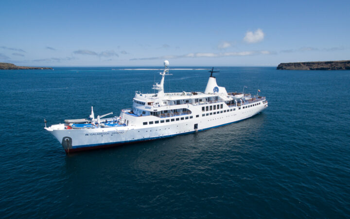 Galapagos Legend cruise ship with white hull & upper decks sailing on blue water photographed from above on a sunny day.