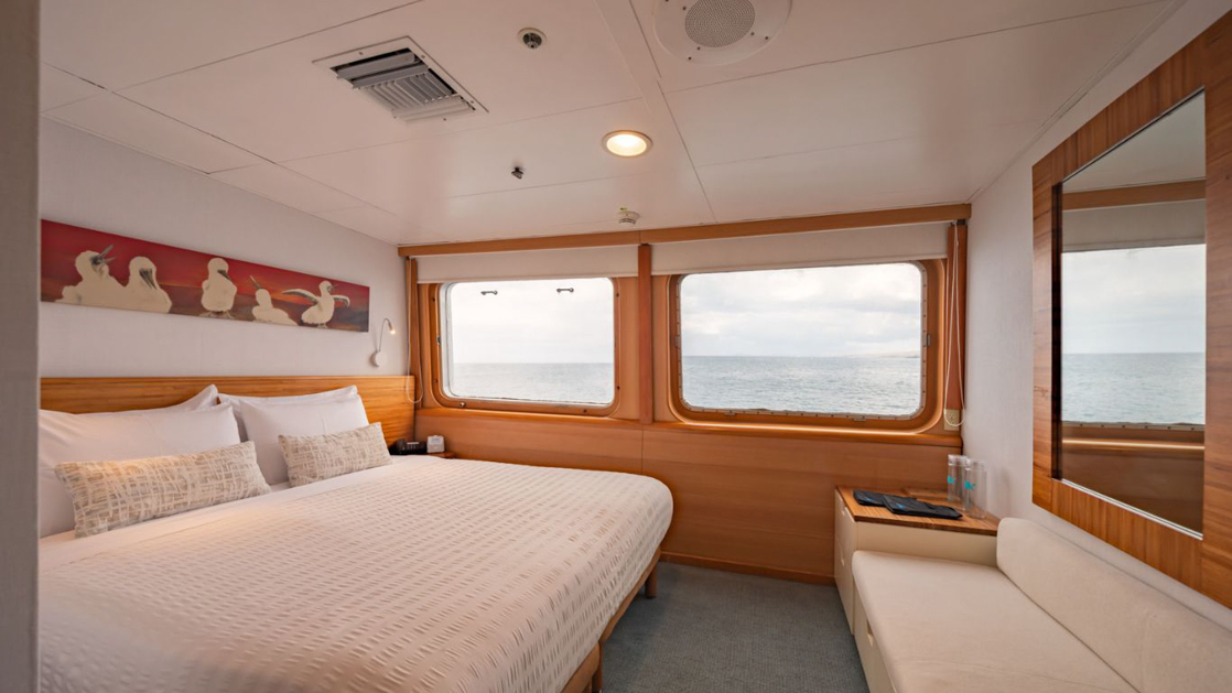 bed aboard small cruise ship with white bedding and a red painting hanging over the bed with large windows looking out into the calm waters