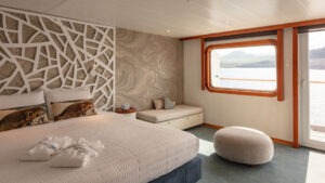 Legend Balcony Suite cabin, with double bed, wraparound sofa, tan-&-green accents, large picture window & private balcony.