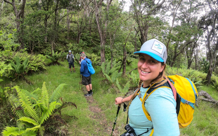 A female traveler in a blue shirt and yellow backpack looks at the camera while hiking on a tropical lush trail in the Hawaiian Islands
